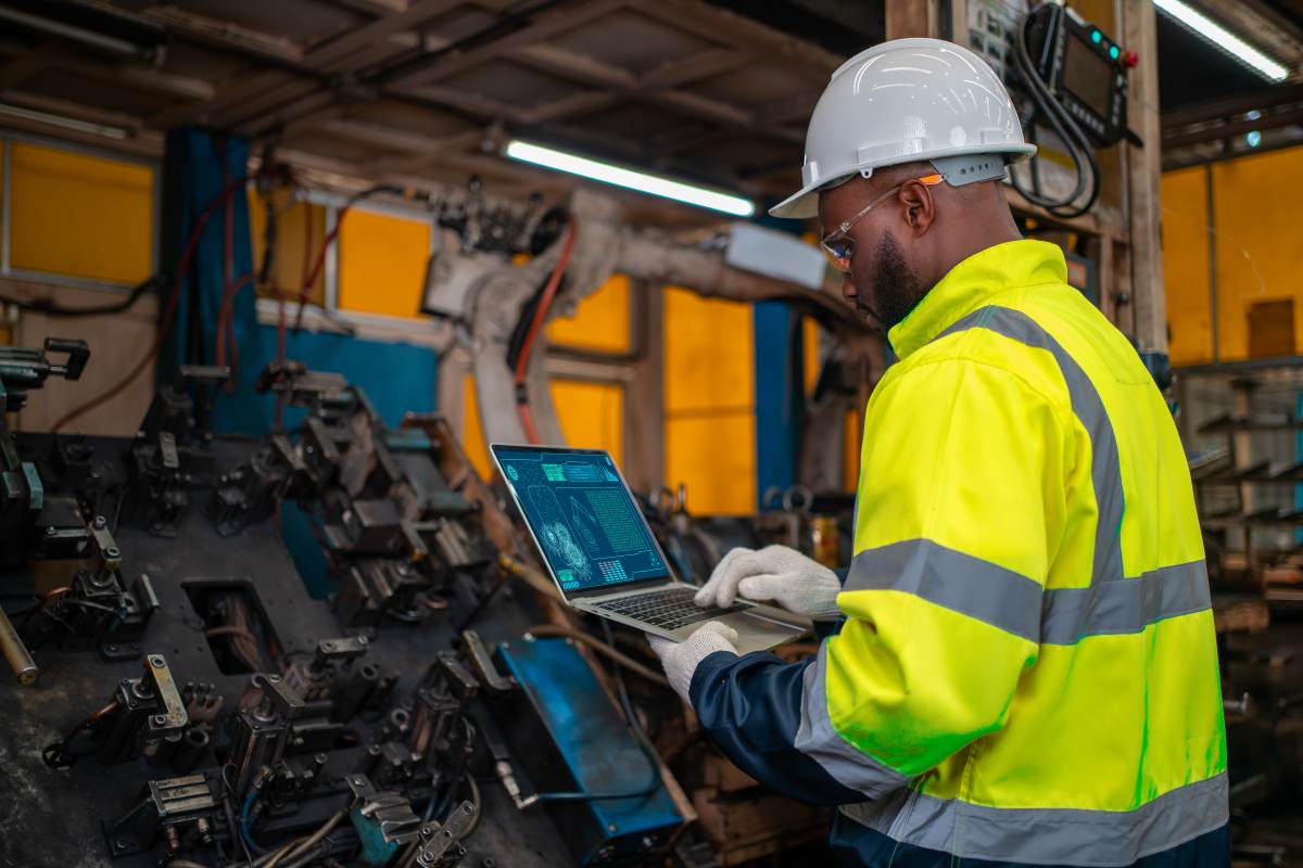 Profissional da indústria com capacete branco e jaleco refletivo amarelo opera um notebook em frente a uma máquina industrial complexa.